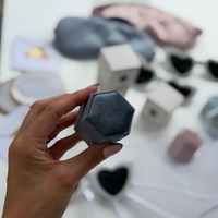 A hand holding a small hexagonal gray velvet jewelry box, with other matching storage boxes and accessories blurred in the background on a white surface.