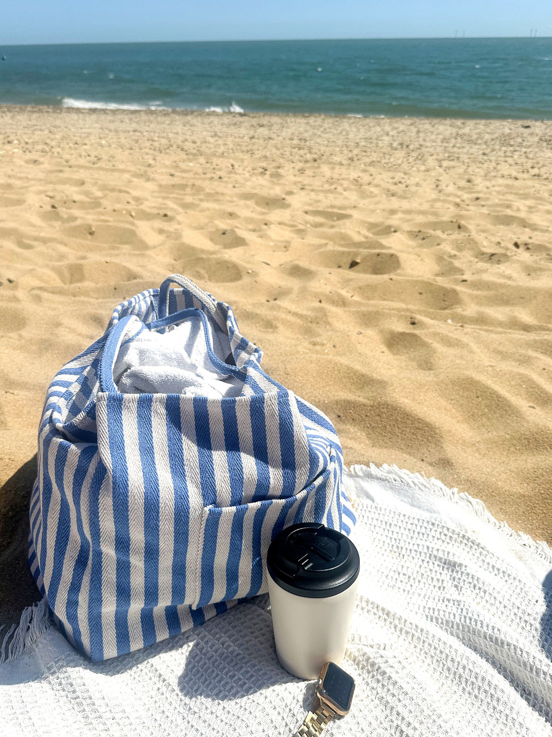 Blue and white striped beach bag and coffee cup on a beach with ocean view